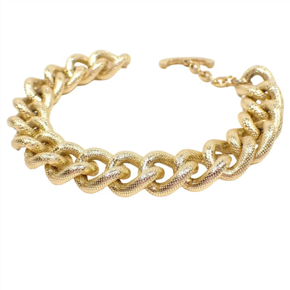 Angled side view of the retro 1975 Avon "Gilded Links" curb chain bracelet. The metal is gold tone in color. The links are wide curved curb shaped and have a light dot textured pattern. There is a toggle clasp at the end.