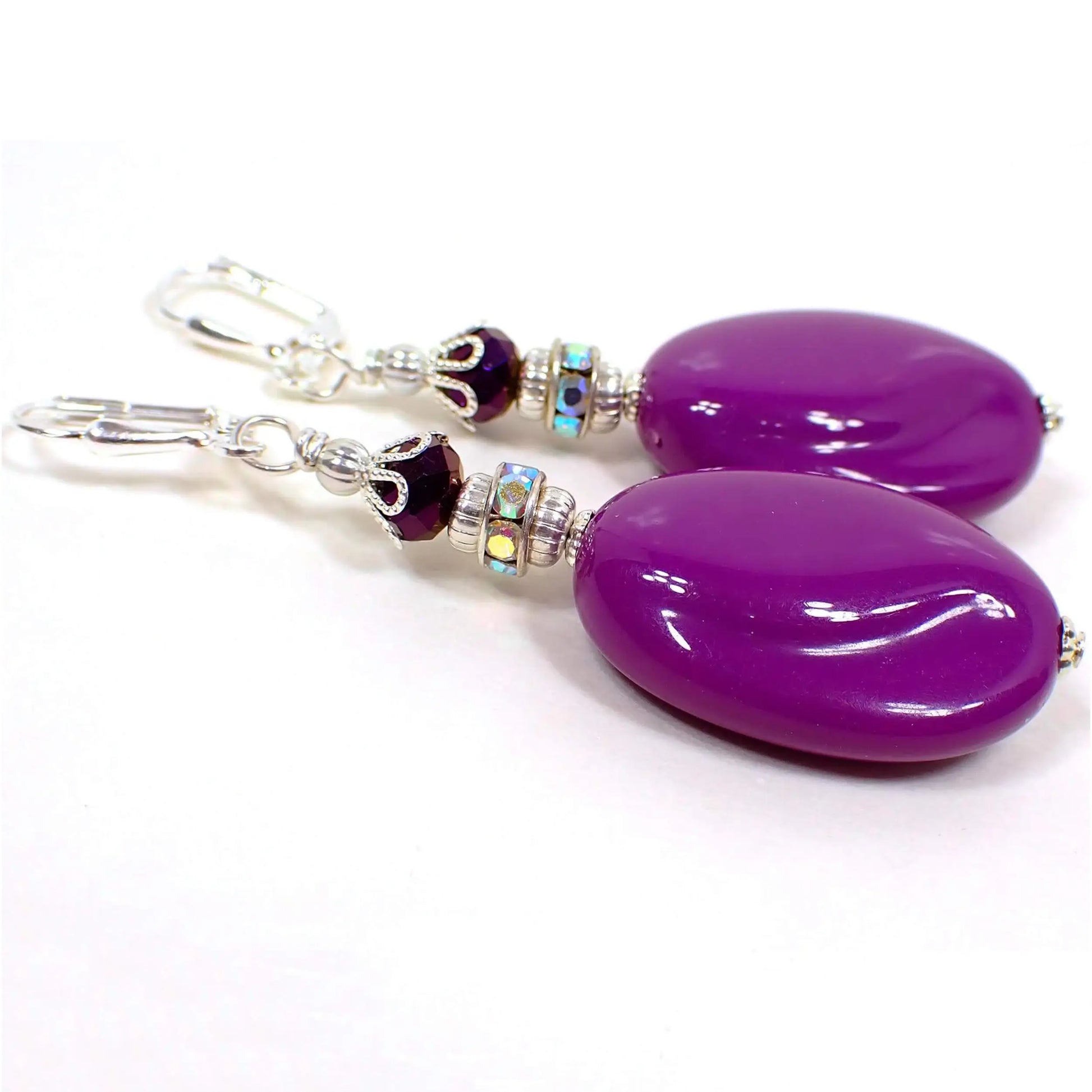 Angled view of the handmade large oval drop earrings made with vintage lucite beads. The metal is silver plated in color. There are metallic purple faceted glass beads at the top. In the middle are beads with AB clear rhinestones all the way around. The bottom lucite beads are a bright purple color and are puffy oval shaped with an indent swirl pattern towards the sides of the beads.