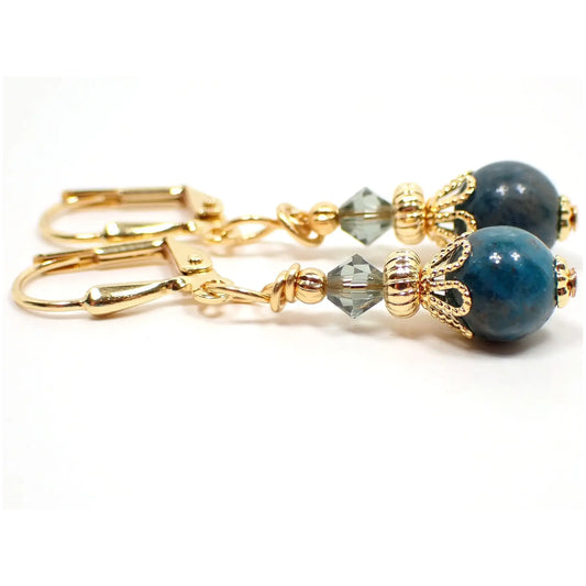 Enlarged side view of the small handmade apatite drop earrings. The metal is gold plated in color. There is a faceted glass crystal light smoky green bead at the top and a small round ball apatite gemstone bead at the bottom. The gemstone is a marbled teal blue in color.