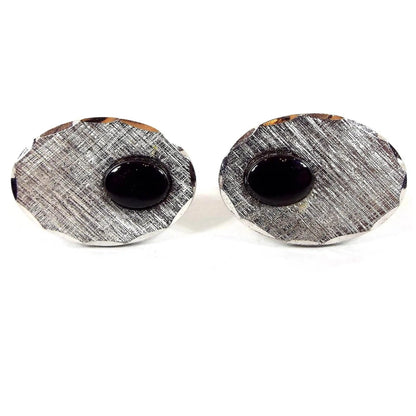 Front view of the JML Mid Century vintage sterling silver and onyx cufflinks. They are oval in shape with a shiny faceted edge and a brushed textured front. There is a small oval black onyx gemstone cab on the side.