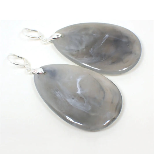 Angled view of the large marbled acrylic teardrop earrings. The metal is silver plated in color. They are large teardrop shaped with shades of gray and small swirls of white marbled in.