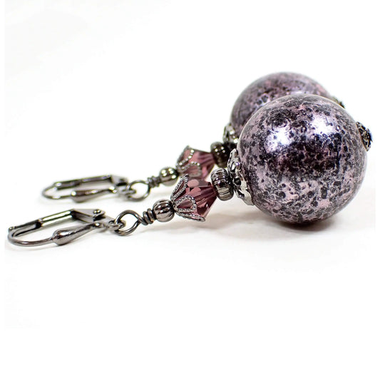 Angled view of the handmade round drop earrings. The metal is gunmetal gray in color. There are purple faceted glass crystal beads at the top. The bottom acrylic beads are sphere ball shaped and are black with light metallic pink and purple brushed on coating.