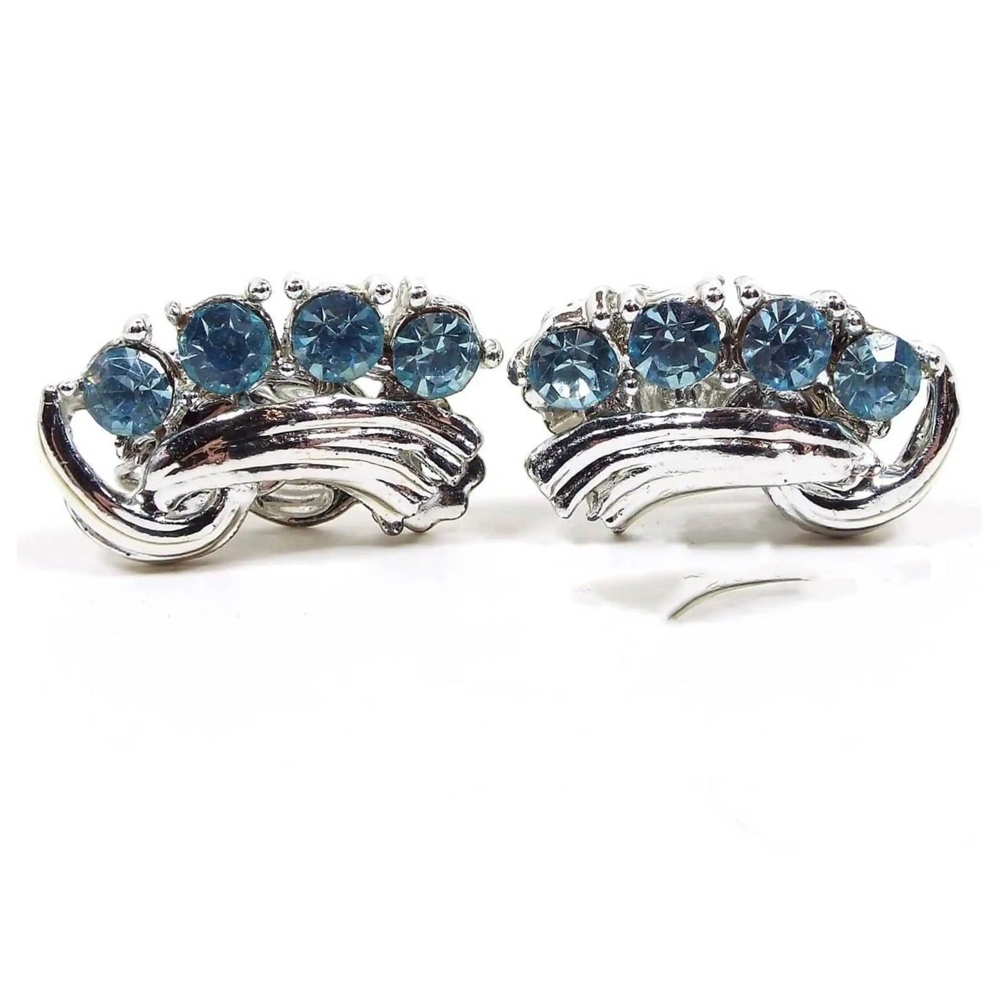 Front view of the Mid Century vintage blue rhinestone earrings. The metal is silver tone in color. There are a row of light blue round rhinestones on one side and a ribbon like strip of silver tone metal on the other.