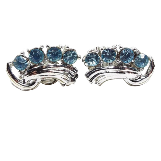 Front view of the Mid Century vintage blue rhinestone earrings. The metal is silver tone in color. There are a row of light blue round rhinestones on one side and a ribbon like strip of silver tone metal on the other.