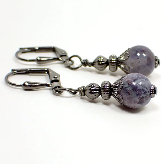 Photo of the small handmade iolite sunstone drop earrings. The metal is gunmetal gray in color. The gemstone beads are small round sphere shaped and are shades of light purple in color with speckles. 