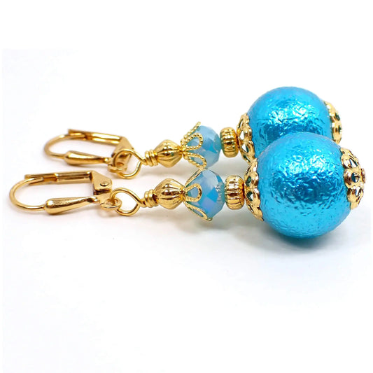 Side view of the handmade sphere drop earrings. The metal is gold plated in color. There is a light blue faceted glass crystal at the top. The bottom acrylic bead is round with a bumpy texture and is a bright aqua blue in color.