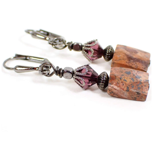 Angled view of the handmade gemstone earrings. The metal is gunmetal gray in color. There are purple faceted glass crystal beads at the top. The bottom jasper beads are small faceted rectangles with shades of brown, reddish brown, and black.
