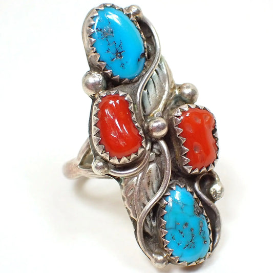 Enlarged front view of the retro vintage Southwestern style ring. The sterling silver has darkened with age to an antiqued silver appearance. There is a an oval turquoise cab at the top with a bean shaped coral cab below it. The ring is divided diagonally by two sterling feathers and then there is another bean shaped coral cab and finally a teardrop shaped turquoise shaped cab at the bottom. There are round sterling balls intermittently around the ring.