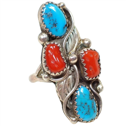 Enlarged front view of the retro vintage Southwestern style ring. The sterling silver has darkened with age to an antiqued silver appearance. There is a an oval turquoise cab at the top with a bean shaped coral cab below it. The ring is divided diagonally by two sterling feathers and then there is another bean shaped coral cab and finally a teardrop shaped turquoise shaped cab at the bottom. There are round sterling balls intermittently around the ring.