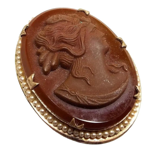 Front view of the Mid Century vintage Jasperware and glass cameo brooch pin. It is oval in shape and has a bust cameo of a woman in Jasperware on top of an oval glass cab. The glass and Jasperware are brown in color. The setting is gold tone in color and has coated imitation round pearls around the edge.