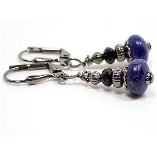 Side view of the small handmade lapis lazuli drop earrings. The metal is gunmetal gray in color. There are black faceted glass crystal beads at the top. The bottom gemstone beads are a dark blue in color with tiny flecks of gold color. The beads are rondelle saucer like shaped.
