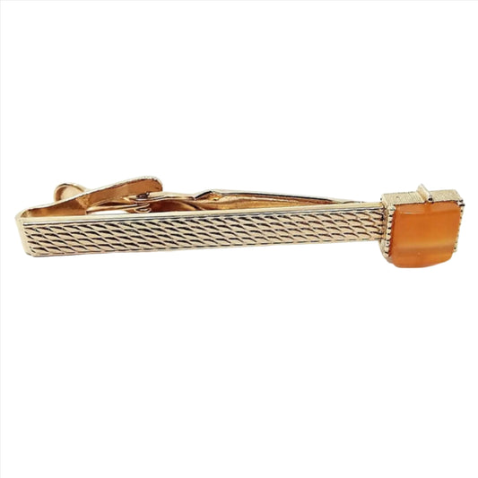Front view of the Mid Century vintage Swank lucite tie clip. The metal is gold tone in color and has a bump scale like pattern on the front. There is a square orange moonglow lucite cab on the end.
