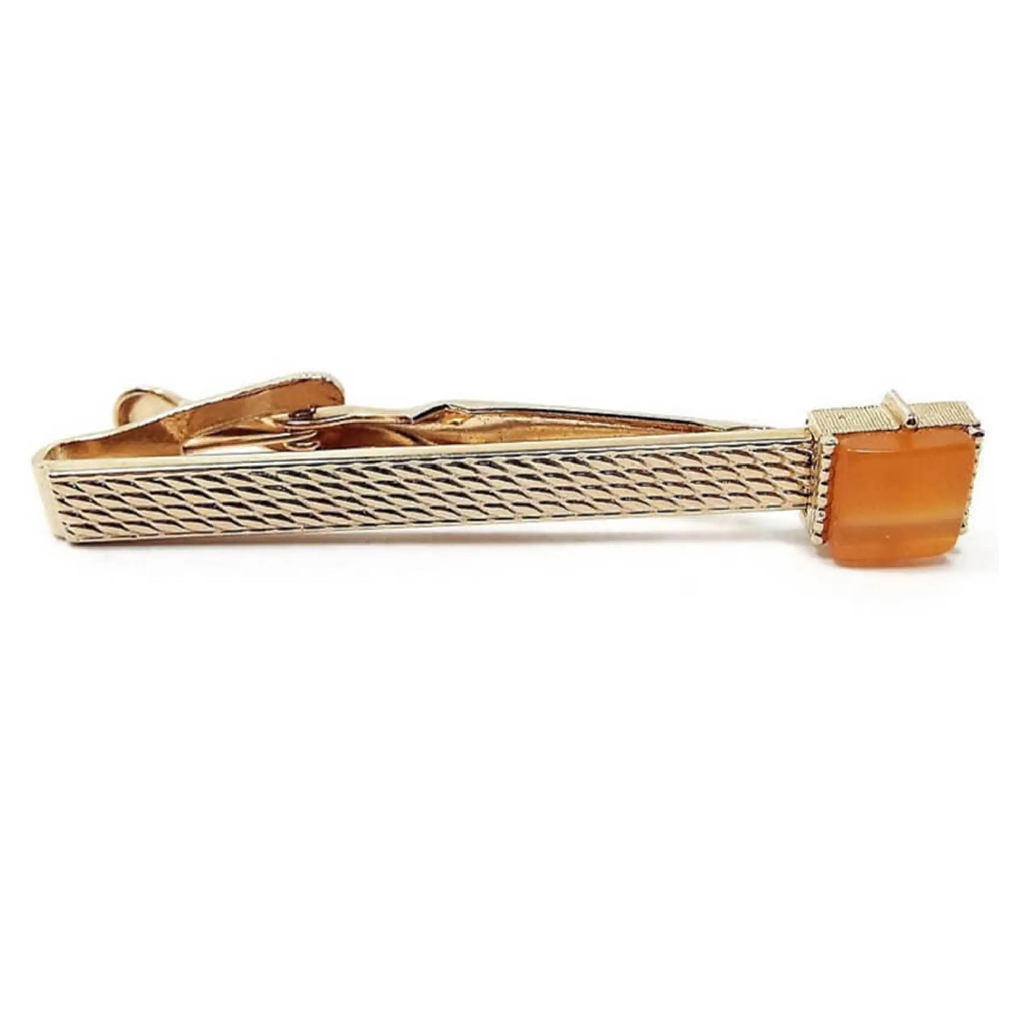 Front view of the Mid Century vintage Swank lucite tie clip. The metal is gold tone in color and has a bump scale like pattern on the front. There is a square orange moonglow lucite cab on the end.