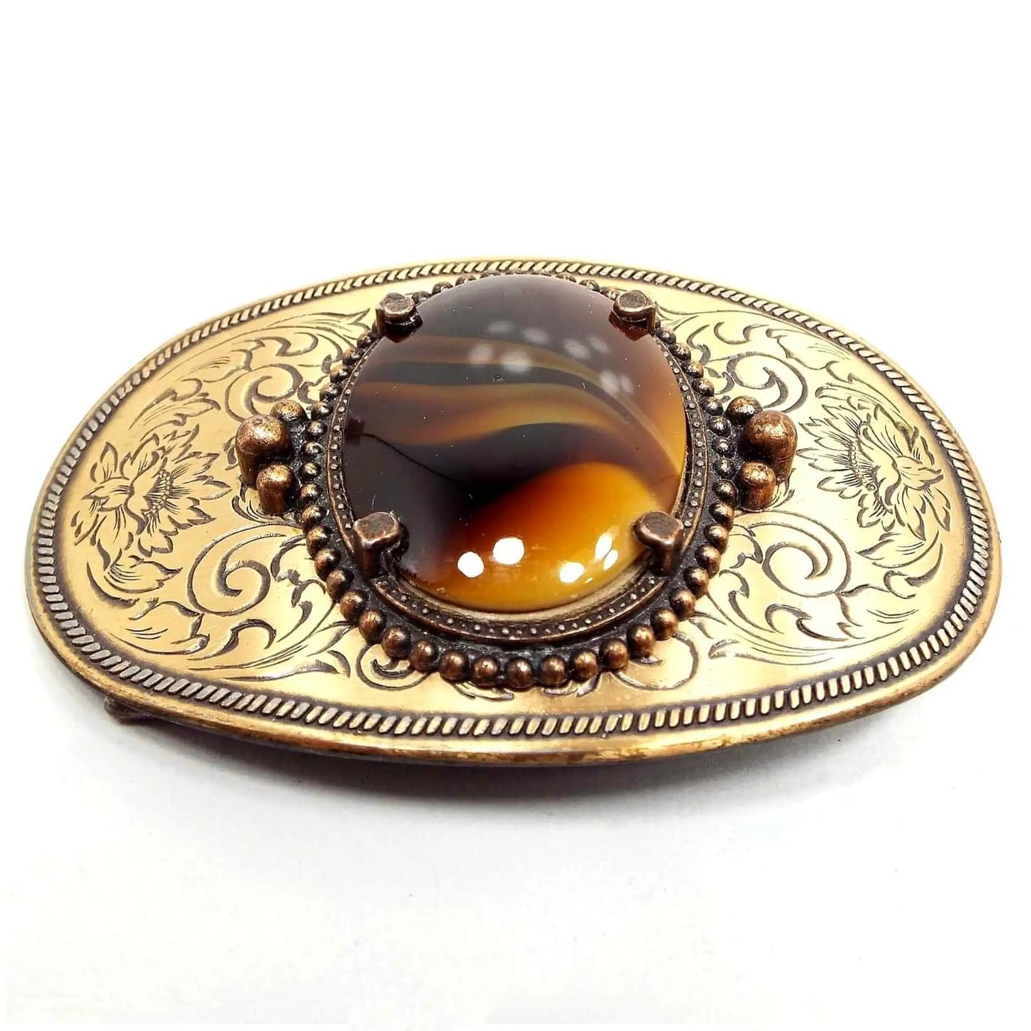 Front view of the retro vintage belt buckle with fancy glass cab. It is oval and has an antiqued gold tone metal color. In the middle is an oval marbled glass cab with shades of brown and yellow. There is an etched floral and leaf design on the metal.