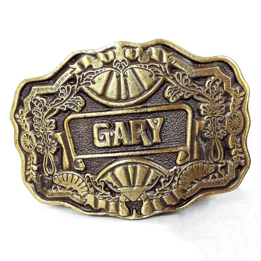 Front view of the retro vintage Gary name belt buckle by Oden. It is antiqued brass in color and has a floral and fan design around the edge. The name Gary is in fancy capital block letters in the middle of the buckle.