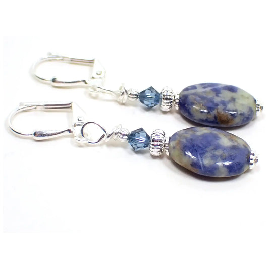Angled view of the handmade sodalite gemstone earrings. The metal is silver plated in color. There are small faceted glass crystal blue beads at the top. The bottom gemstone beads are puffy oval shaped and are denim blue with marbled areas that have shades of cream and brown colors.