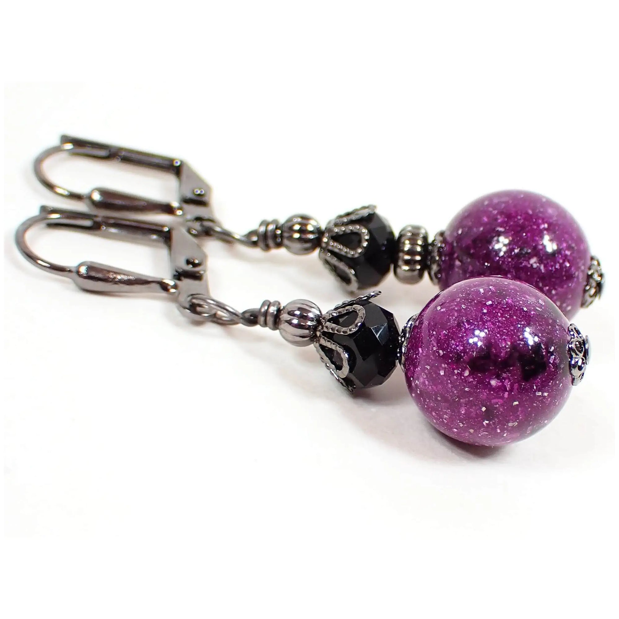 Angled view of the handmade drop earrings made with vintage lucite beads. The metal is gunmetal gray in color. There is a faceted black crystal glass bead at the top. The bottom lucite beads are round ball shaped and are purple with marbled areas of black and tiny flecks of confetti and glitter for a bit of sparkle as you move around in the light.