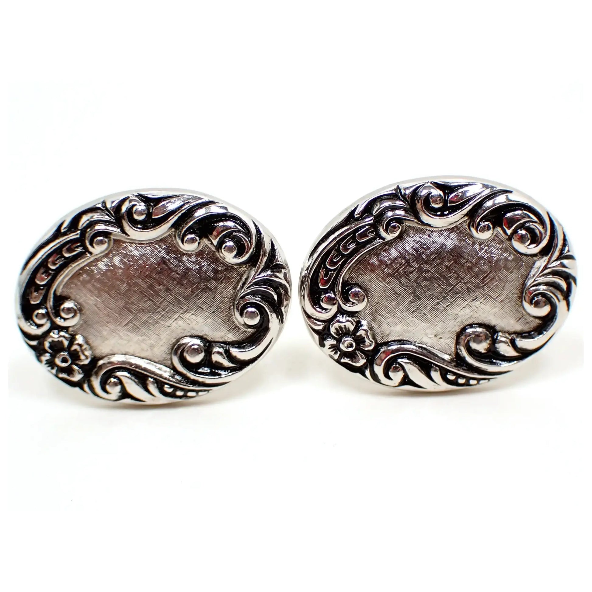 Enlarged front view of the Mid Century vintage Sarah Coventry cufflinks. The metal is antiqued silver tone in color. They have a brush textured front on the oval shapes, and have a scroll design with curls and a small flower around the edge.