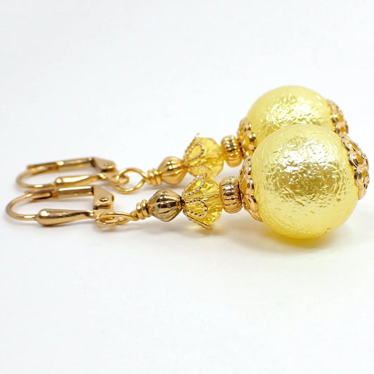 Side view of the handmade sphere drop earrings. The metal is gold plated in color. There is a light yellow faceted glass crystal at the top. The bottom acrylic bead is round with a bumpy texture and is also light yellow in color.