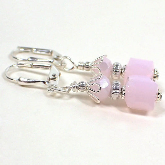 Side view of the handmade small cube drop earrings. The metal is silver plated in color. There are faceted glass rondelle beads at the top and square cube beads at the bottom in a light opaque pink color.