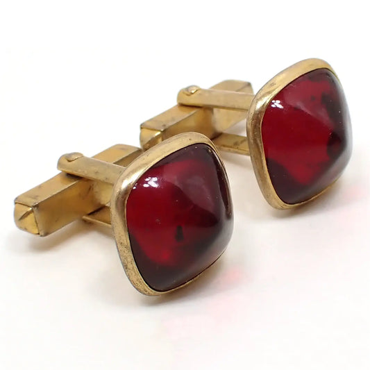 Enlarged angled photo of the Mid Century vintage Swank lucite cufflinks. The metal is antiqued brass in color. They are shaped like rounded squares and have domed dark red lucite fronts.