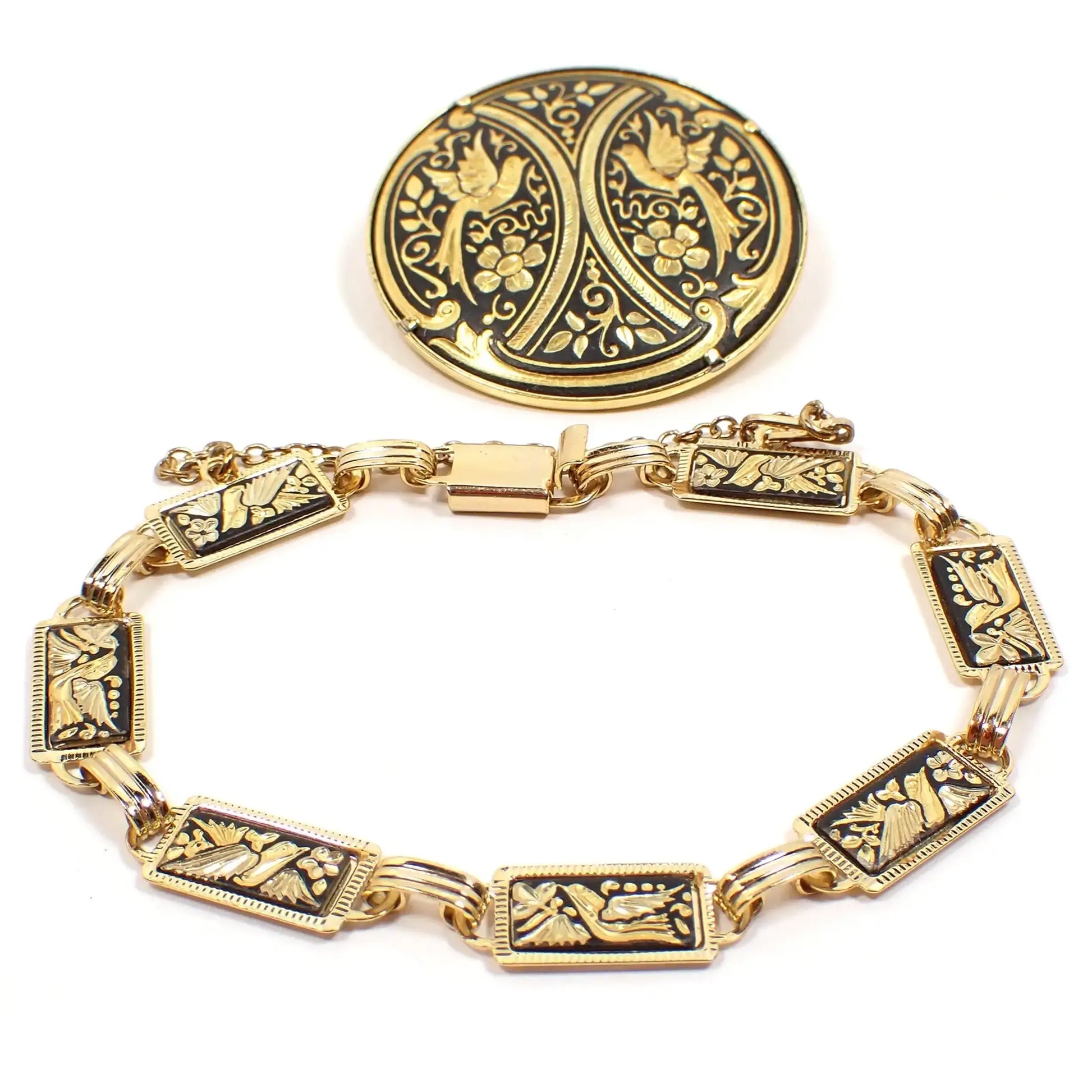 Photo of retro vintage Midas of Spain Damascene jewelry set. There is a link bracelet at the bottom with gold tone metal. The links are black with birds and flowers etched in different shades of gold tone metal. There is a box clasp at the end. Above the bracelet is a matching round brooch with bird and floral design in black and gold color as well.