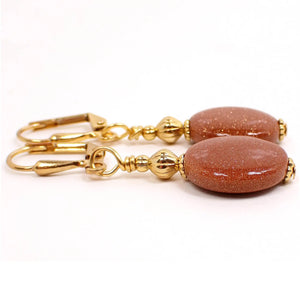 Side view of the handmade Goldstone earrings. The metal is gold plated in color. There are two metal beads at the top and a flat oval goldstone bead at the bottom. The goldstone bead is glass with very tiny specks of copper glitter. The earrings sparkle and glisten as you move around in the light.