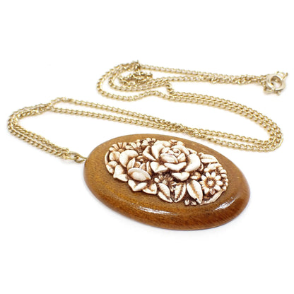 Angled enlarged front view of the retro vintage floral cameo pendant necklace. The chain is gold tone in color with a spring ring clasp at the end. There is a wooden oval pendant at the bottom with a plastic floral cameo cab design on the front. The flowers are off white in color with brown in the recessed areas.