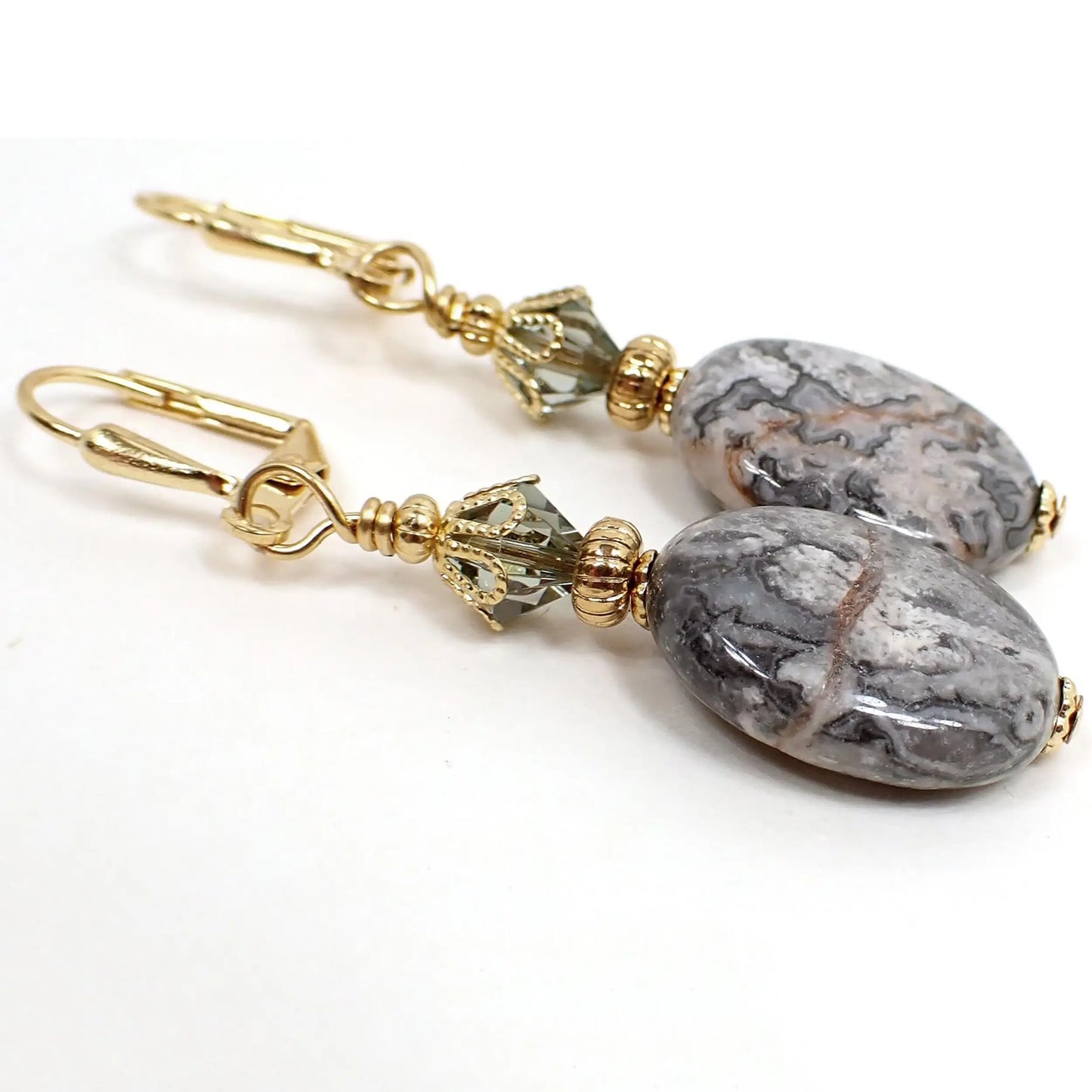 Angled view of the handmade crazy lace agate gemstone earrings. The metal is gold plated in color. There are faceted glass crystal beads at the top in a smoky greenish gray color. The bottom gemstone beads are oval shaped and have mostly marbled gray colors with a few red lines through them here and there.