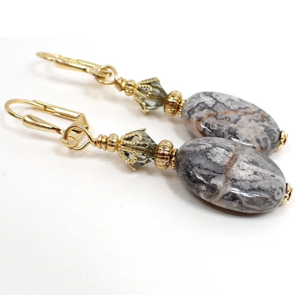 Angled view of the handmade crazy lace agate gemstone earrings. The metal is gold plated in color. There are faceted glass crystal beads at the top in a smoky greenish gray color. The bottom gemstone beads are oval shaped and have mostly marbled gray colors with a few red lines through them here and there.