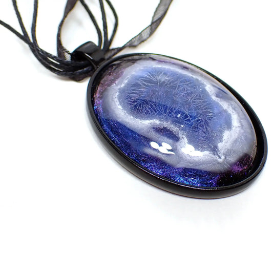 Enlarged front view of the handmade Goth large oval pendant necklace. The necklace part shows three strands of black faux leather cord and a strand of black organza ribbon. The pendant is oval in shape with a black coated base metal setting. The cab is made of resin with an iridescent blue background that has hints of purple at the top and bottom in the photo. There is a white frost like crystalline design in the middle with an abstract edge.