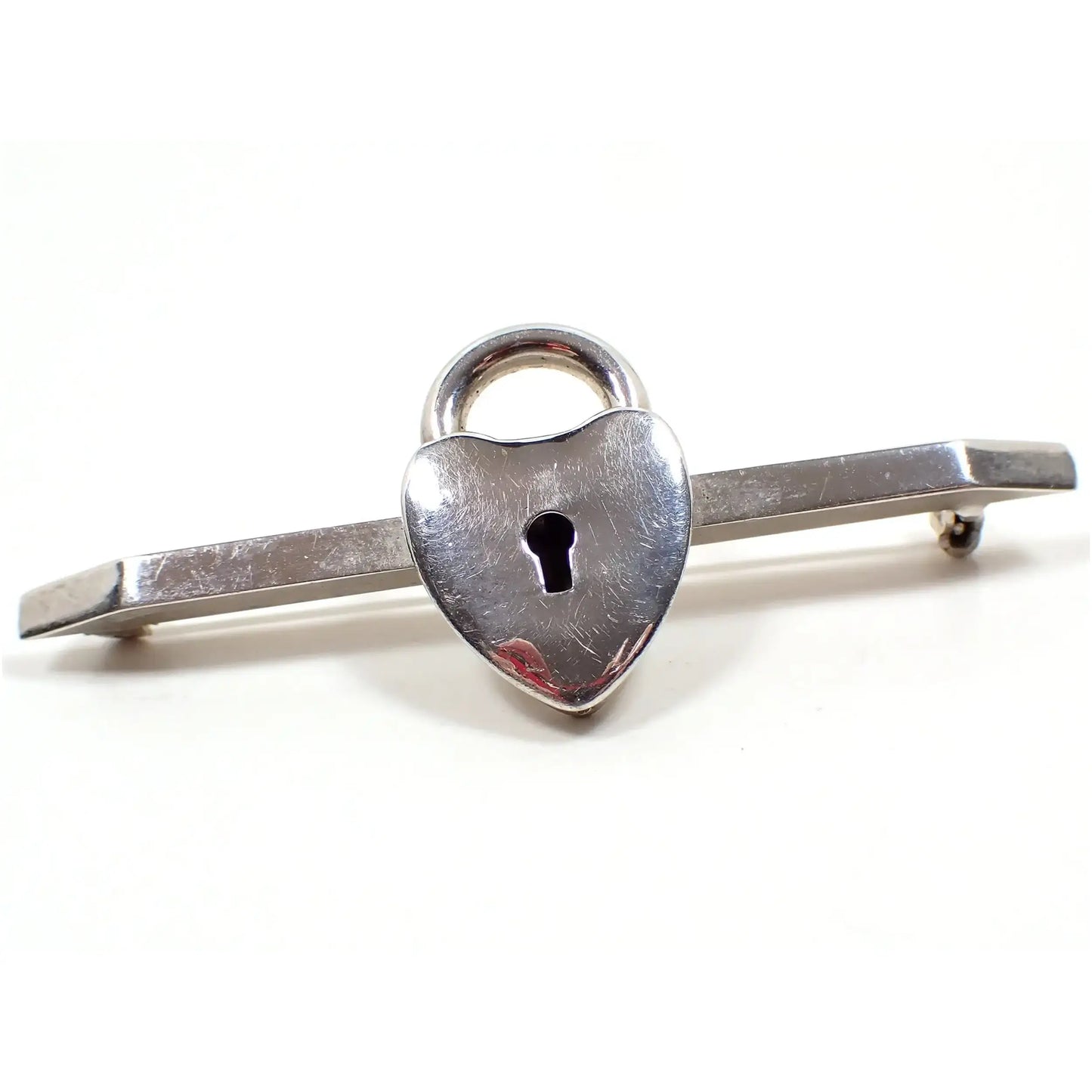 Enlarged view of the retro vintage padlock style bar brooch. The bar is angled on the ends and the metal is silver tone in color. In the middle is a fake lock shaped like a heart that has a skeleton key hold in the middle.