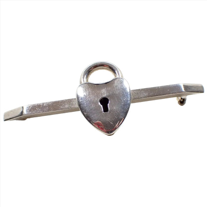 Enlarged view of the retro vintage padlock style bar brooch. The bar is angled on the ends and the metal is silver tone in color. In the middle is a fake lock shaped like a heart that has a skeleton key hold in the middle.