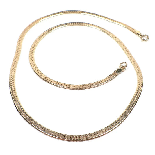 Top view of the retro vintage Krementz herringbone chain necklace. It is gold tone in color and has a flat herringbone link design. There is a spring ring clasp at one end and a ring at the other end that has markings on it.
