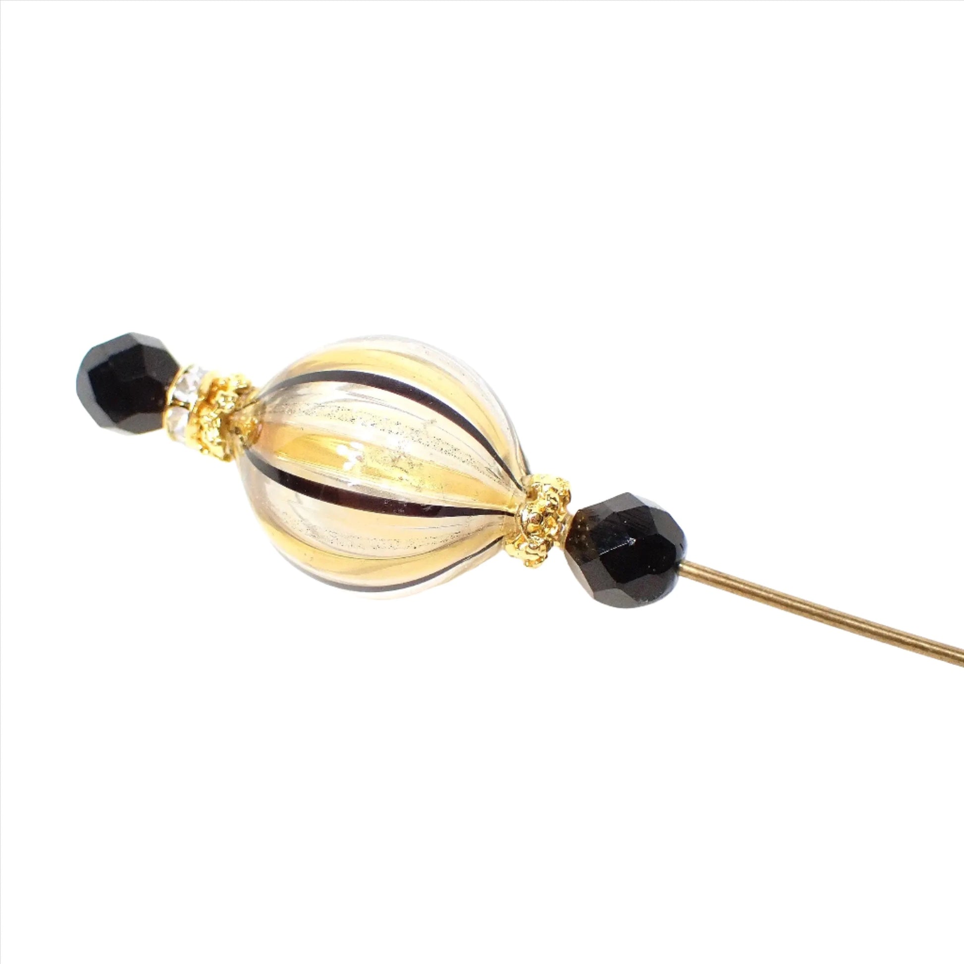 Enlarged top view of the Mid Century vintage hat pin with Murano Glass top. The metal is gold tone plated in color. There is a round hollow Murano glass ball at the top that has black and gold color stripes with fine metallic glitter in between. There is a faceted black bead and rhinestone bead on top of the Murano glass and another faceted black glass bead below it. There is a slide on pin clutch at the bottom.