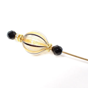 Enlarged top view of the Mid Century vintage hat pin with Murano Glass top. The metal is gold tone plated in color. There is a round hollow Murano glass ball at the top that has black and gold color stripes with fine metallic glitter in between. There is a faceted black bead and rhinestone bead on top of the Murano glass and another faceted black glass bead below it. There is a slide on pin clutch at the bottom.