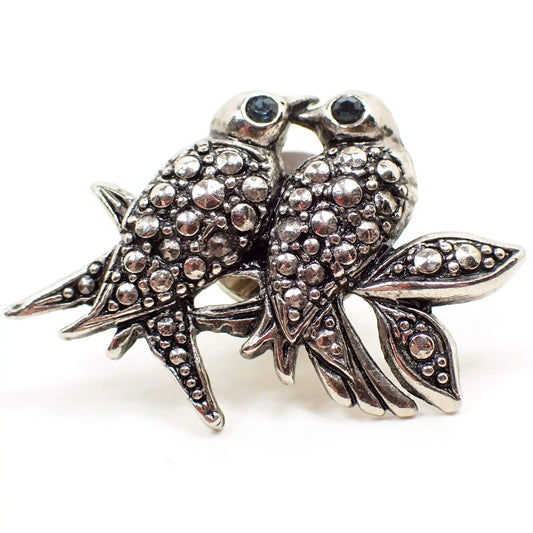 Enlarged front view of the retro vintage birds tie tack. The metal is antiqued silver tone plated in color. There are two birds sitting on a branch with two leaves on the right. The birds and leaves have an imitation steel cut design of rounded dots. Each bird has a blue rhinestone eye.