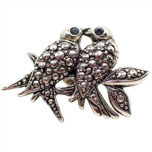Enlarged front view of the retro vintage birds tie tack. The metal is antiqued silver tone plated in color. There are two birds sitting on a branch with two leaves on the right. The birds and leaves have an imitation steel cut design of rounded dots. Each bird has a blue rhinestone eye.