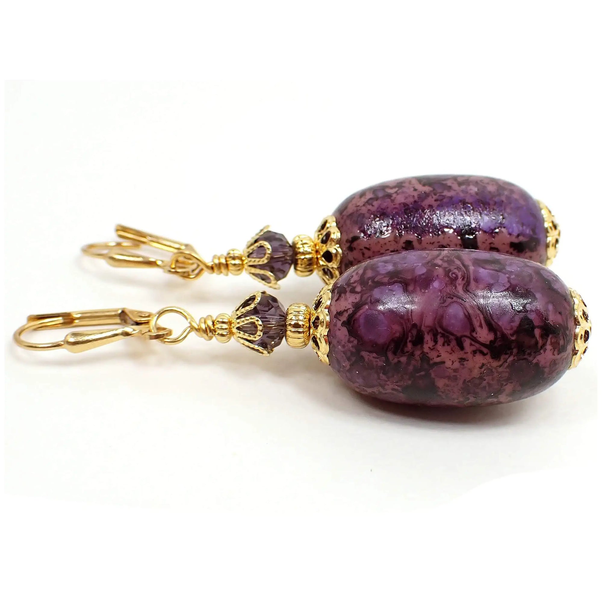 Side view of the large oval handmade drop earrings. The metal is gold plated in color. There are purple faceted glass crystal beads at the top. The bottom acrylic beads are large oval shaped and have a marbled pattern with purple and pink.