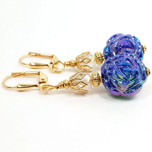 top view of the handmade flower earrings with AB blue acrylic beads. The metal is gold plated in color. The top beads are AB clear faceted ovals. The bottom beads are shaped like flowers and are primarily blue with AB coating that gives flashes of other colors like pink and green and purple as you move around in the light.