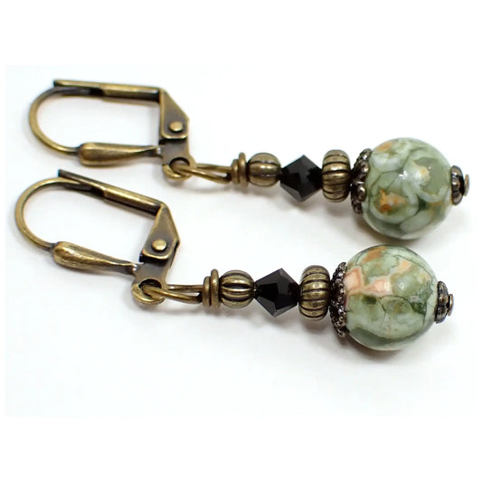 Enlarged view of the handmade rainforest jasper earrings. The metal is antiqued brass in color. There are small faceted black crystal bicone beads at the top. The bottom gemstone beads are round sphere shaped and have marbled shades of green, peach, and white.