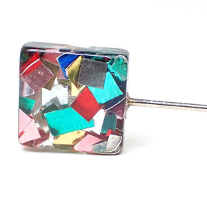 Enlarged view of the top of the pre owned stick pin. It is square in shape with clear resin. Embedded in the rest are square and rectangular shaped pieces of colored thin metal for a rainbow multi color appearance.