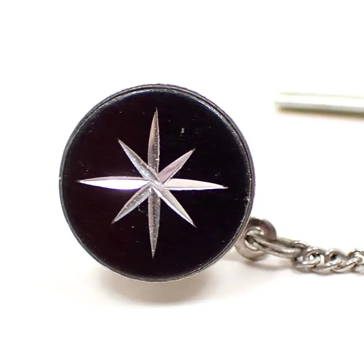 Enlarged front view of the Mid Century vintage tie tack. It is round with a black painted front. There is an engraved atomic starburst design in the middle that shows the silver plated metal underneath.