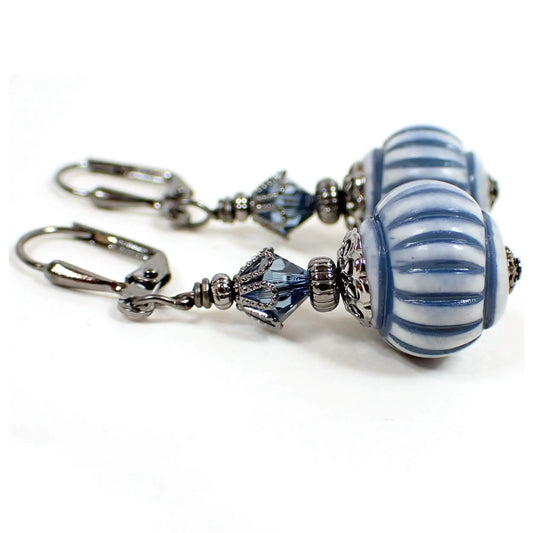Side view of the handmade lantern drop earrings. The metal is gunmetal gray in color. There are blue faceted glass crystal beads at the top. The bottom beads are acrylic and are lantern shaped round. They are light blue in color with stripes in a country blue color.