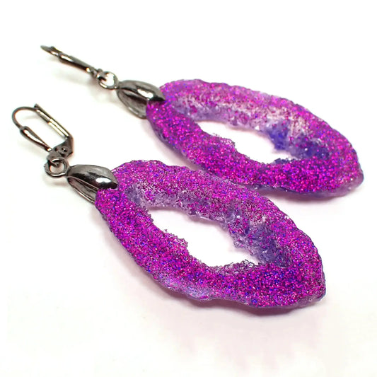Angled front view of the handmade faux druzy geode slice earrings. The metal is dark gunmetal gray in color. The drops are long oval shaped with an open middle and druzy like edges. The earrings have purple resin and are loaded with lots of bright fuchsia pink purple iridescent glitter on the front.