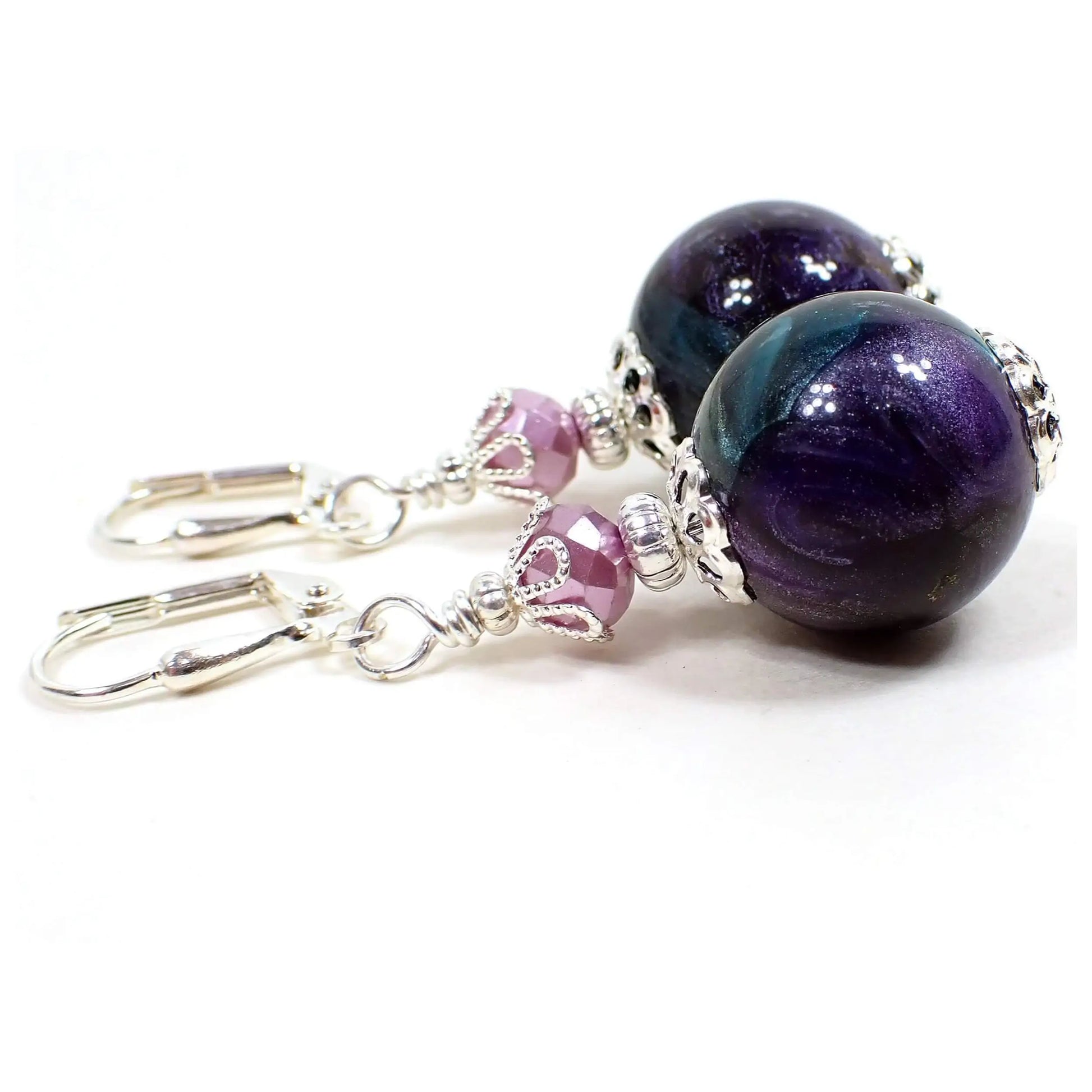 Angled view of the handmade multi color earrings. The metal is silver plated in color. There are new opaque faceted glass beads in a light purple. The bottom beads are vintage lucite and are round ball shaped and have pearly marbled swirls of different colors such as blue, green, and purple.