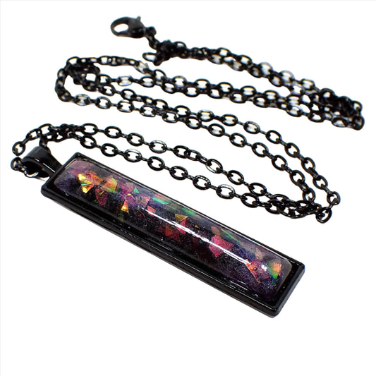 Enlarged front view of the handmade resin bar necklace. The metal is coated black in color. The front resin cab has dark pearly purple resin with chunks of iridescent glitter that flashes different colors as it moves around in the light.