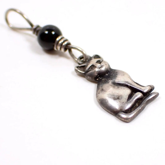 Enlarged angled view of the small retro vintage cat charm. The charm is shaped like a sitting cat. There is a wire wrapped round ball onyx gemstone bead attached to the top of the charm.