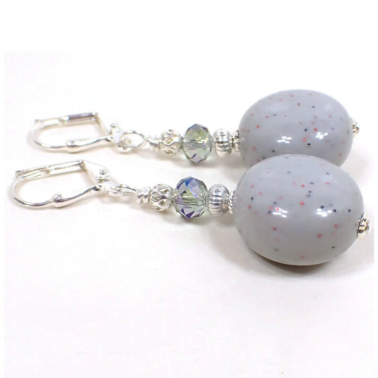 Angled view of the handmade drop earrings. The metal is silver plated in color. There are faceted blue and gray glass crystal beads at the top. The bottom beads are reclaimed vintage confetti lucite beads and are a light bluish gray in color with tiny flecks of blue and pink confetti glitter embedded in them. They are a puffy round shape.