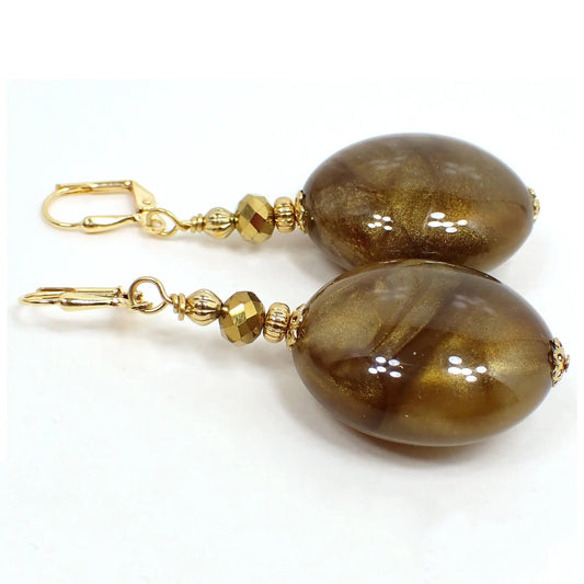 Angled view of the big oval handmade drop earrings with vintage lucite beads. The metal is gold plated in color. There are faceted metallic bronze glass beads at the top. The bottom beads are large puffy ovals that have marbled swirls in different shades of pearly golden brown.